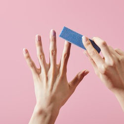 Hand buffing natural nails with a blue 4-way nail buffer against a pink background